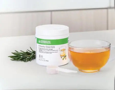 La imagen muestra un envase blanco de Herbalife Pro, un producto para fortalecer el sistema inmune. En el frente del envase está el logo de Herbalife y el nombre del producto en letras grandes. El fondo es completamente blanco. tambien aparece una taza con un liquido