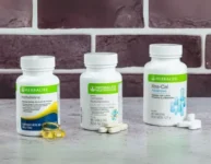 Foto de una variedad de Vitaminas y Suplementos Herbalife, incluyendo frascos de multivitaminas, calcio, y suplementos específicos para el corazon como omega 3 todos exhibidos en un fondo de pared ede ladrillo que resalta la calidad y presentación de los productos.